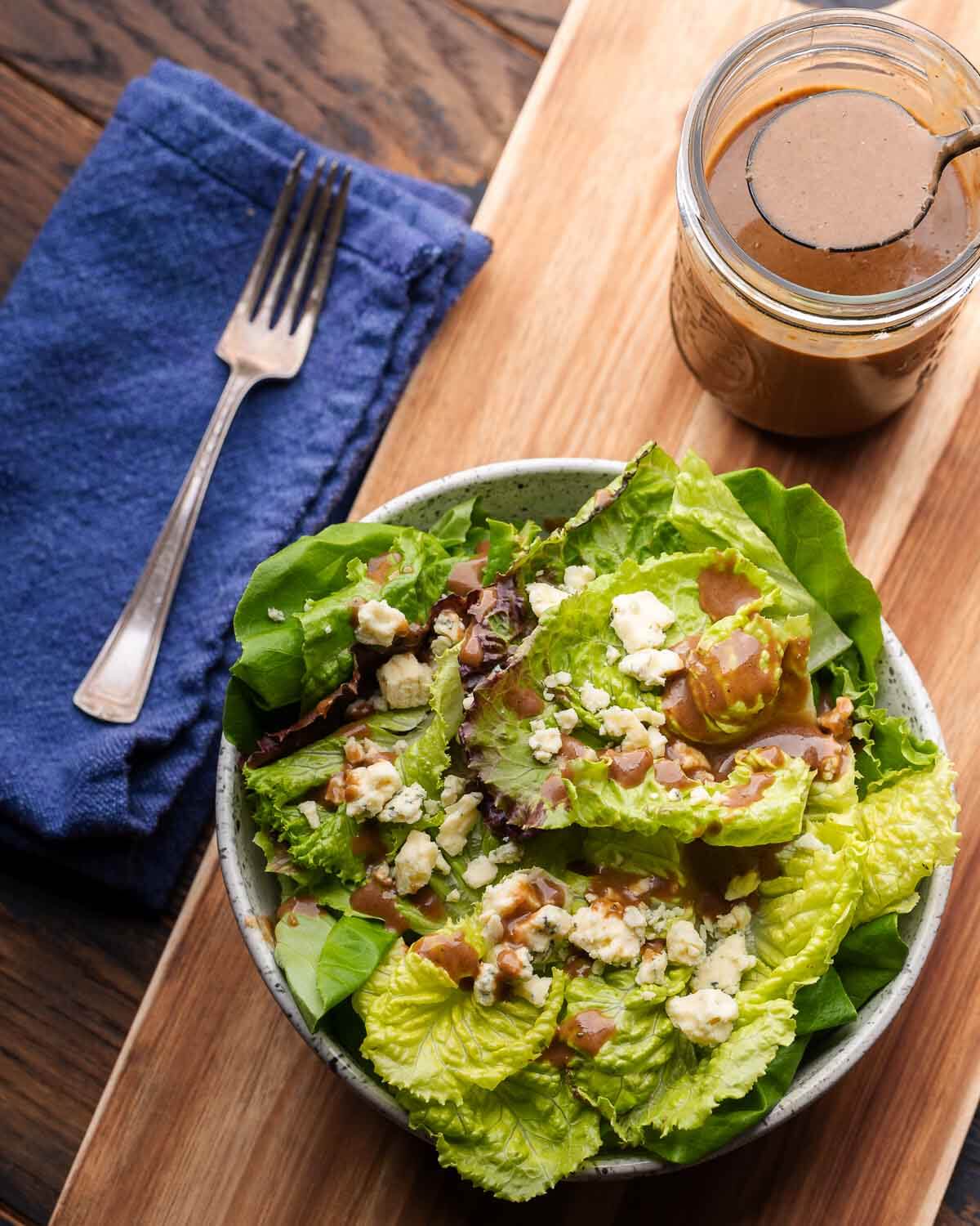 Bowl of salad, fork on blue napkin, and mason jar filled with balsamic vinaigrette.