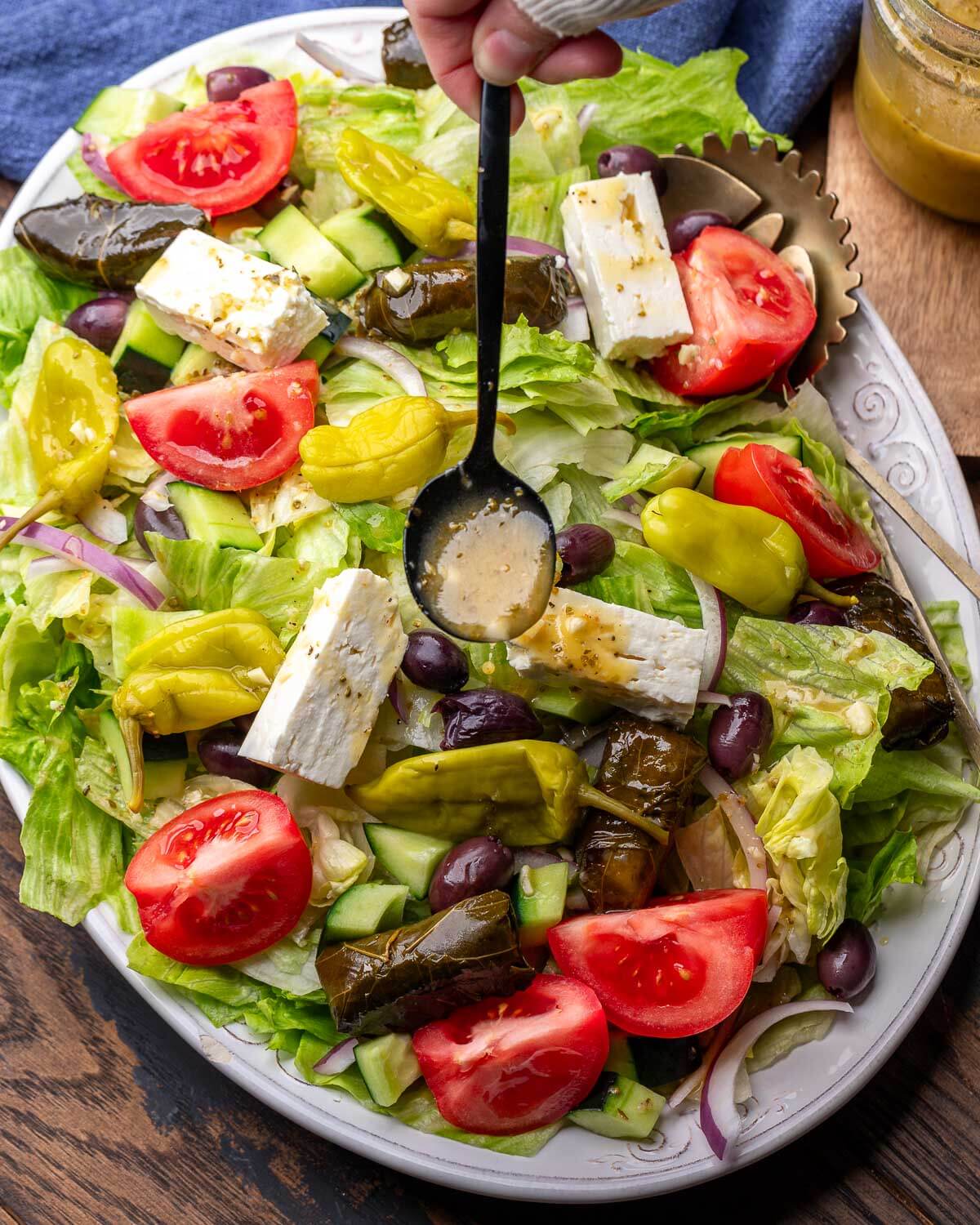 Diner-style Greek salad in white platter with spoon drizzling Greek vinaigrette on top.