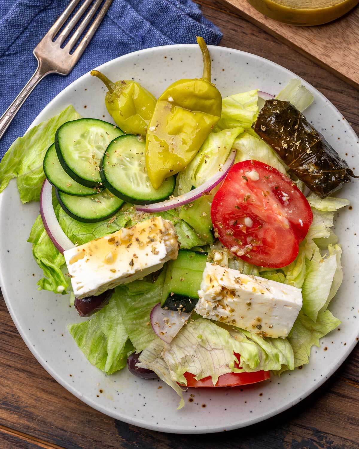 White plate with Greek salad topped with feta cheese, Tuscan peppers, grape leaves, cucumbers, and tomatoes.