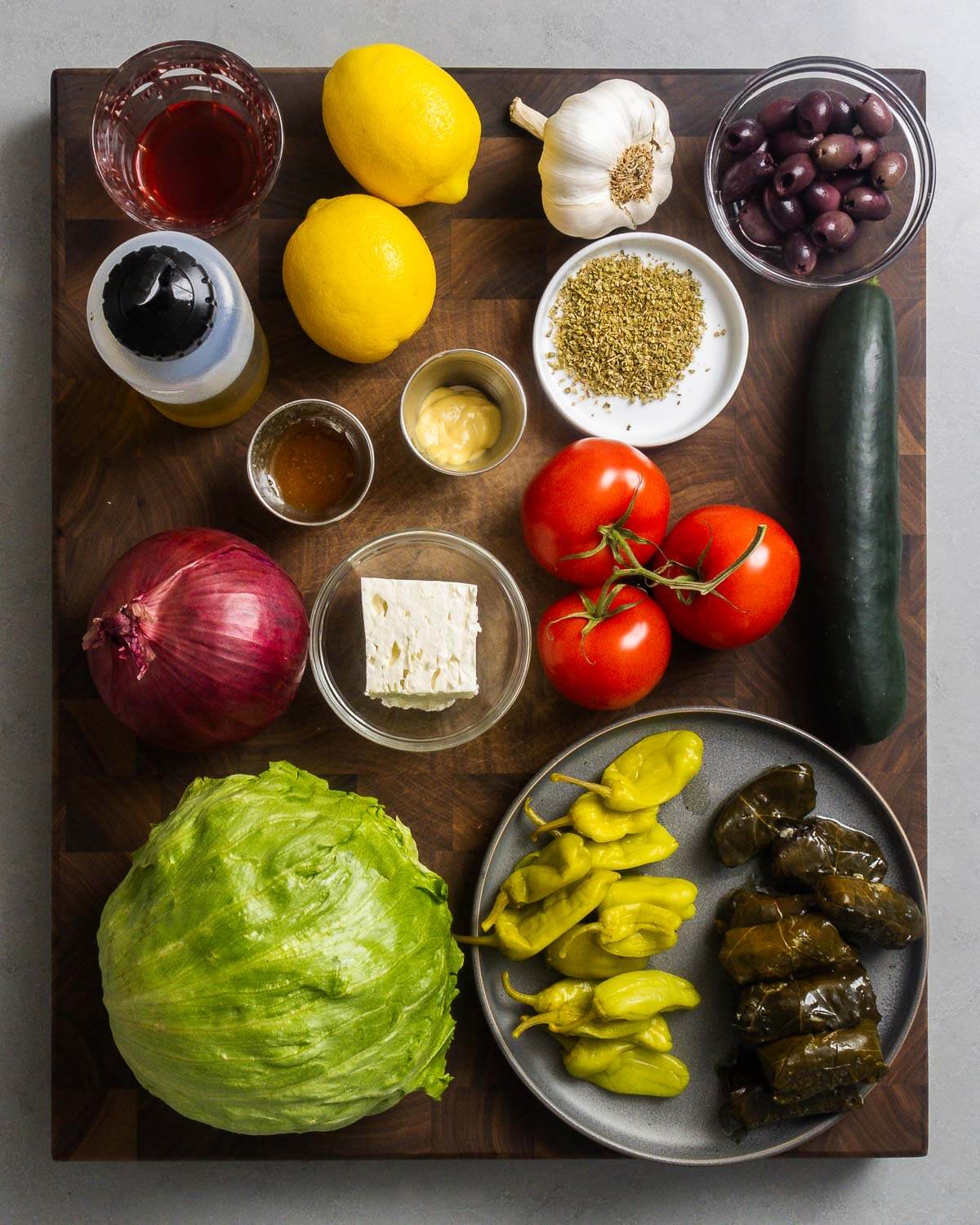 Ingredients shown: red wine vinegar, lemons, olive oil, garlic, kalmata olives, oregano, honey, Dijon mustard, onion, feta cheese, tomatoes, cucumber, iceberg lettuce, Tuscan peppers, and stuffed grape leaves.