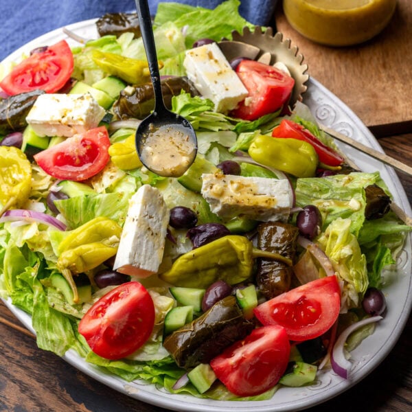 Diner Greek salad on large white platter with spoon drizzling Greek vinaigrette on top.