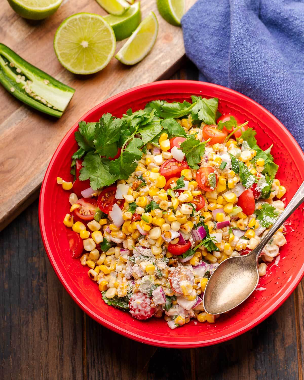Red bowl of street corn salad with blue napkin and limes and jalapenos on cutting board.
