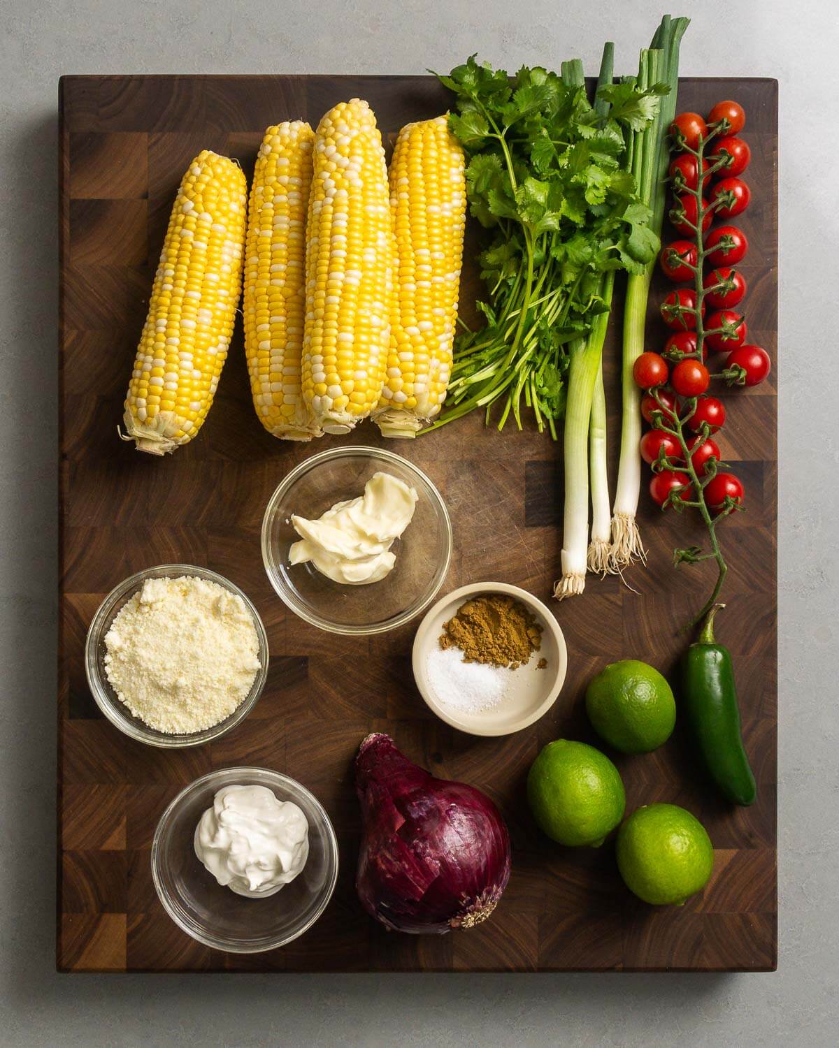 Ingredients shown: corn cobs, cilantro, green onion, cherry tomatoes, cotija cheese, mayonnaise, salt, cumin, sour cream, red onion, limes, and jalapeno pepper.