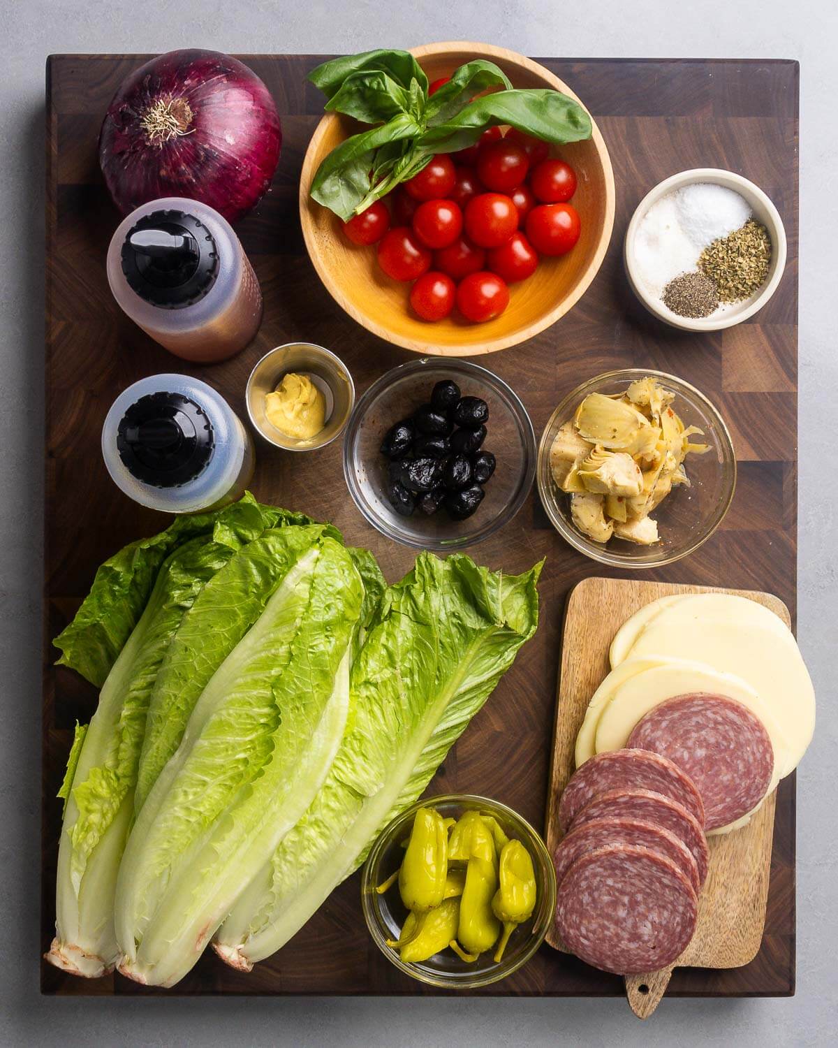 Ingredients shown: red onion, basil, tomatoes, spices, red wine vinegar, olive oil, Dijon mustard, olives, artichoke hearts, lettuce, pepperoncini, provolone, and salami.