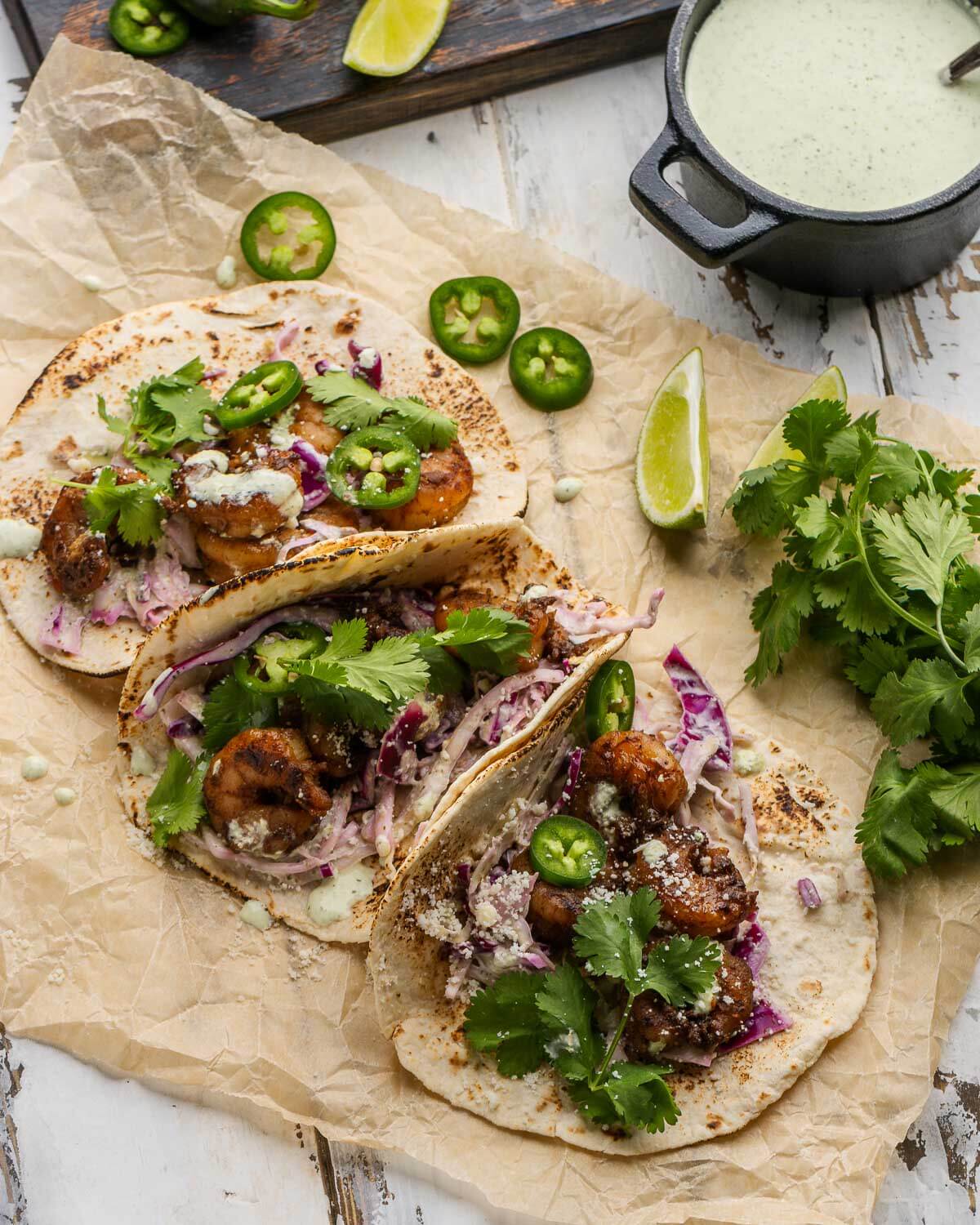 Three shrimp tacos on parchment paper with jalapenos, lime wedges, and cilantro along with black bowl of jalapeno cream sauce.