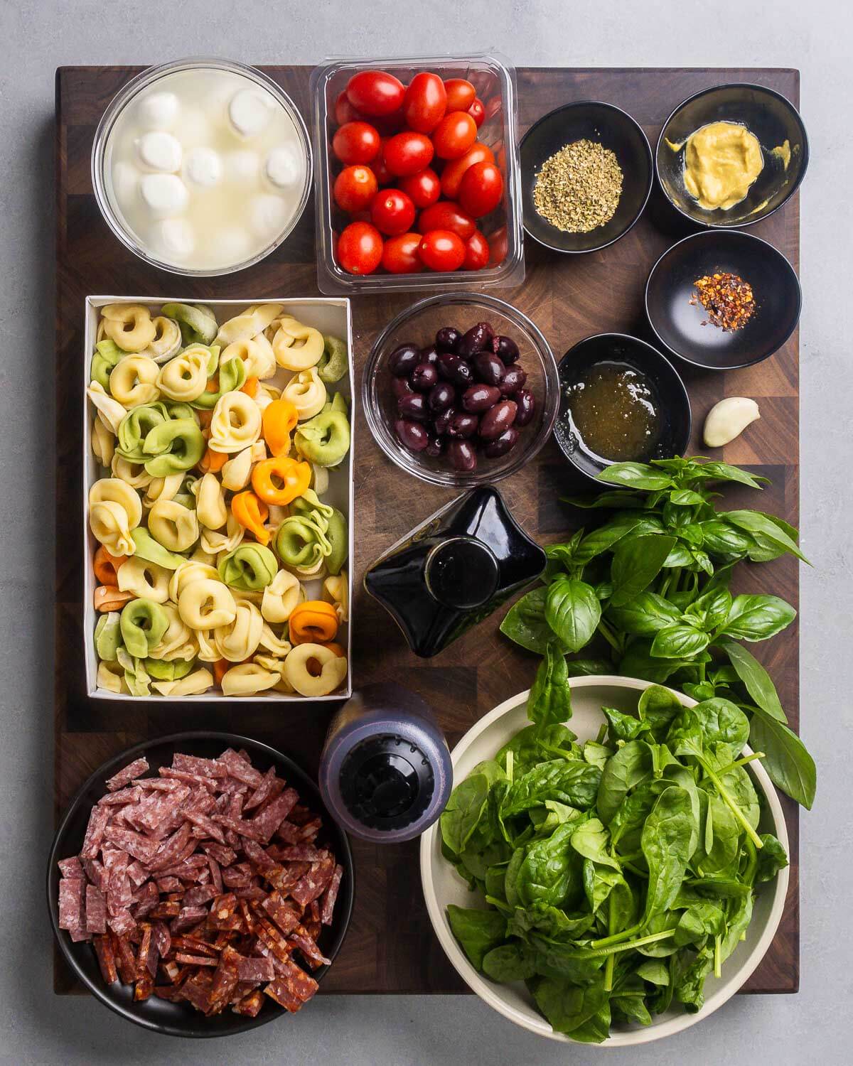 Ingredients shown: mozzarella balls, tomatoes, spices, olives, tortellini, olive oil, basil, red wine vinegar, salami, soppressata, and spinach.