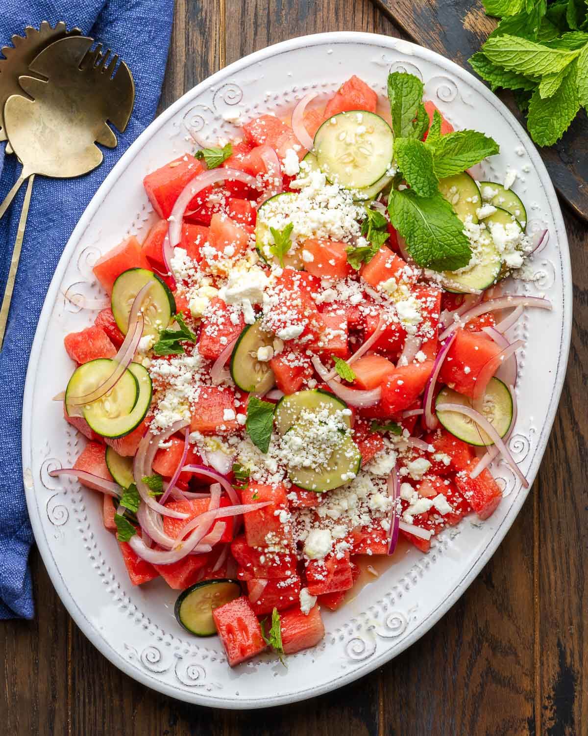 White platter with watermelon feta salad on wood table along with bronze serving spoons and mint on cutting board.