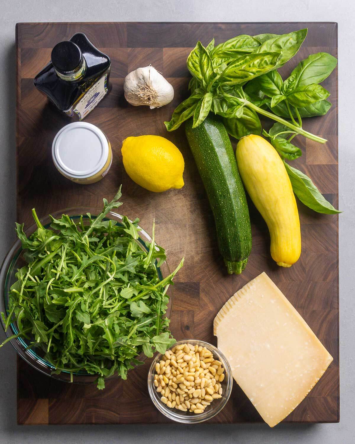 Ingredients shown: extra virgin olive oil, garlic, Dijon mustard, lemon, basil, zucchini, yellow squash, baby arugula, pine nuts, and block of Parmigiano Reggiano cheese.