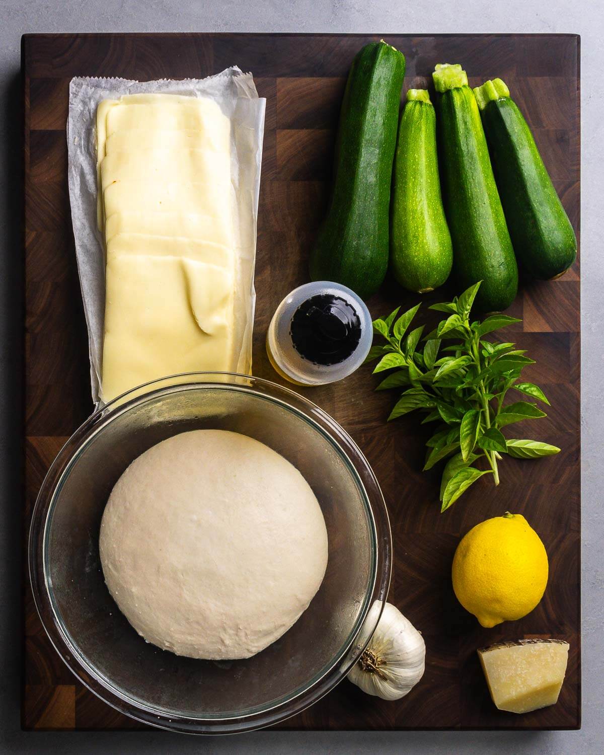 Ingredients shown: sliced mozzarella cheese, zucchini, olive oil, basil, pizza dough, garlic, lemon, and Pecorino Romano cheese.
