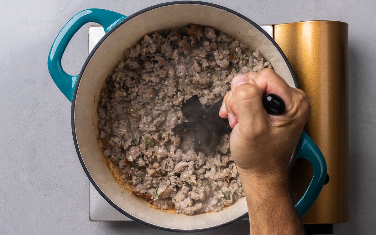 A hand holding a meat masher over pot with sausage.