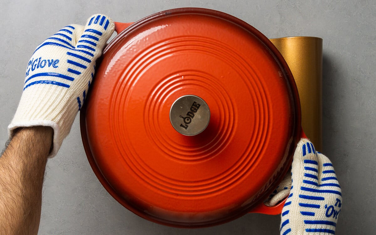 Two hands wearing oven gloves holding an orange dutch oven.