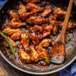 Black pan with balsamic braised chicken wings and rosemary garnish and wooden spoon.