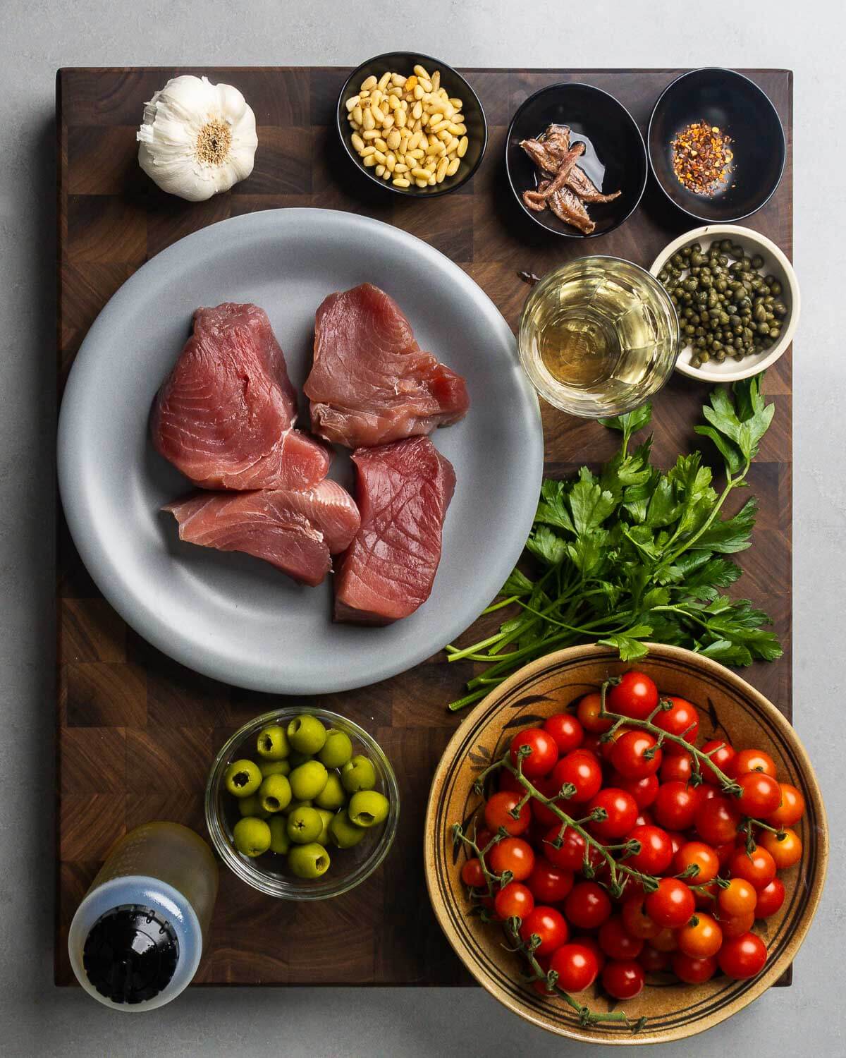 Ingredients shown: garlic, pignoli nuts, anchovies, hot red pepper, wine, capers, tuna steaks, parsley, olive oil, olives, and cherry tomatoes.