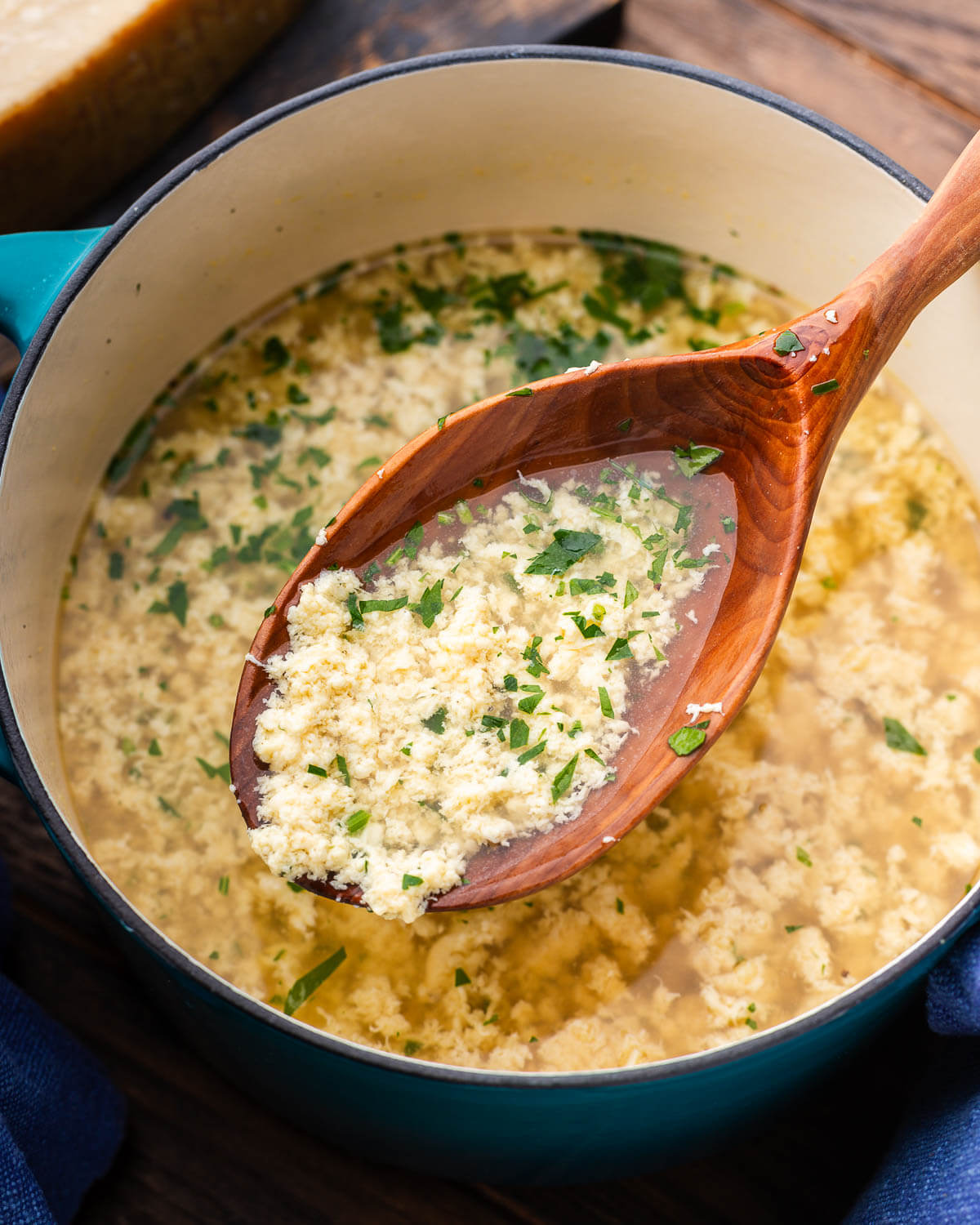Ladle of stracciatella soup held over pot with block of parmesan in the background.