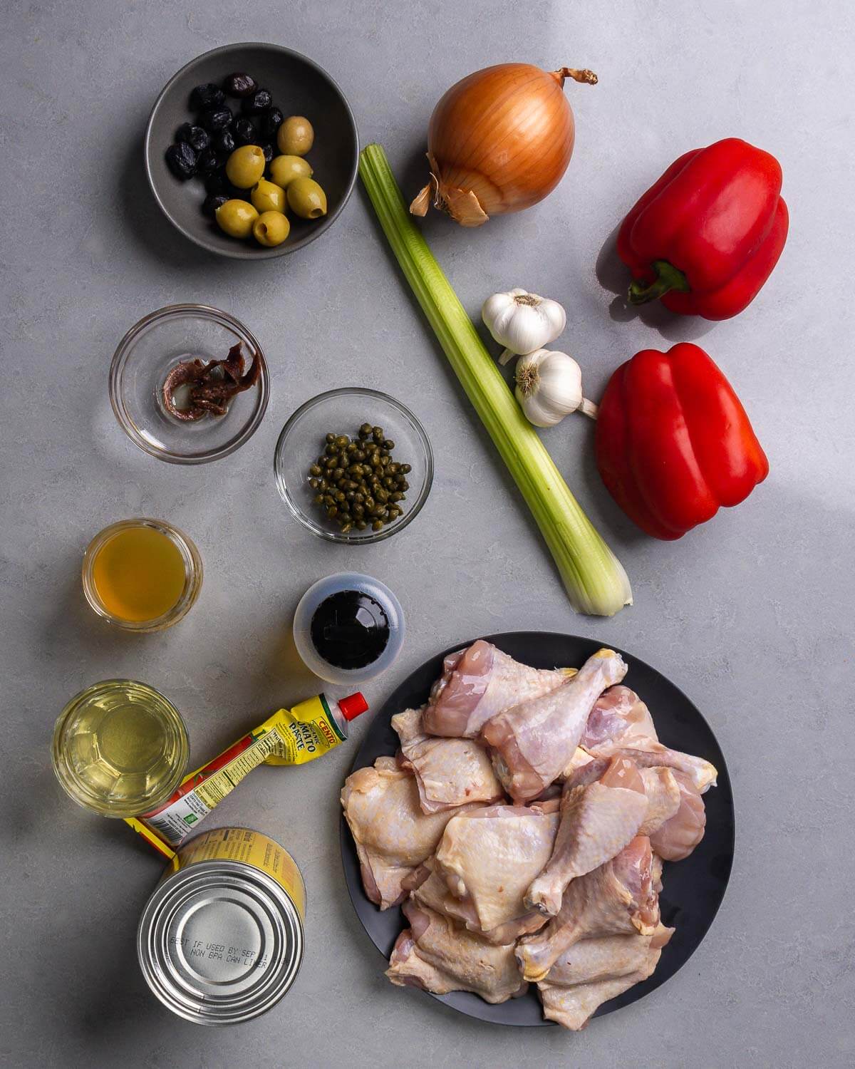 Ingredients shown: olives, onion, celery, garlic, bell peppers, anchovies, capers, wine, chicken stock, olive oil, tomato paste, canned tomatoes, and chicken thighs and legs.