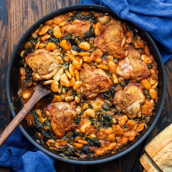 Chicken thighs with brothy beans in large black pan with loaf of bread on the side.