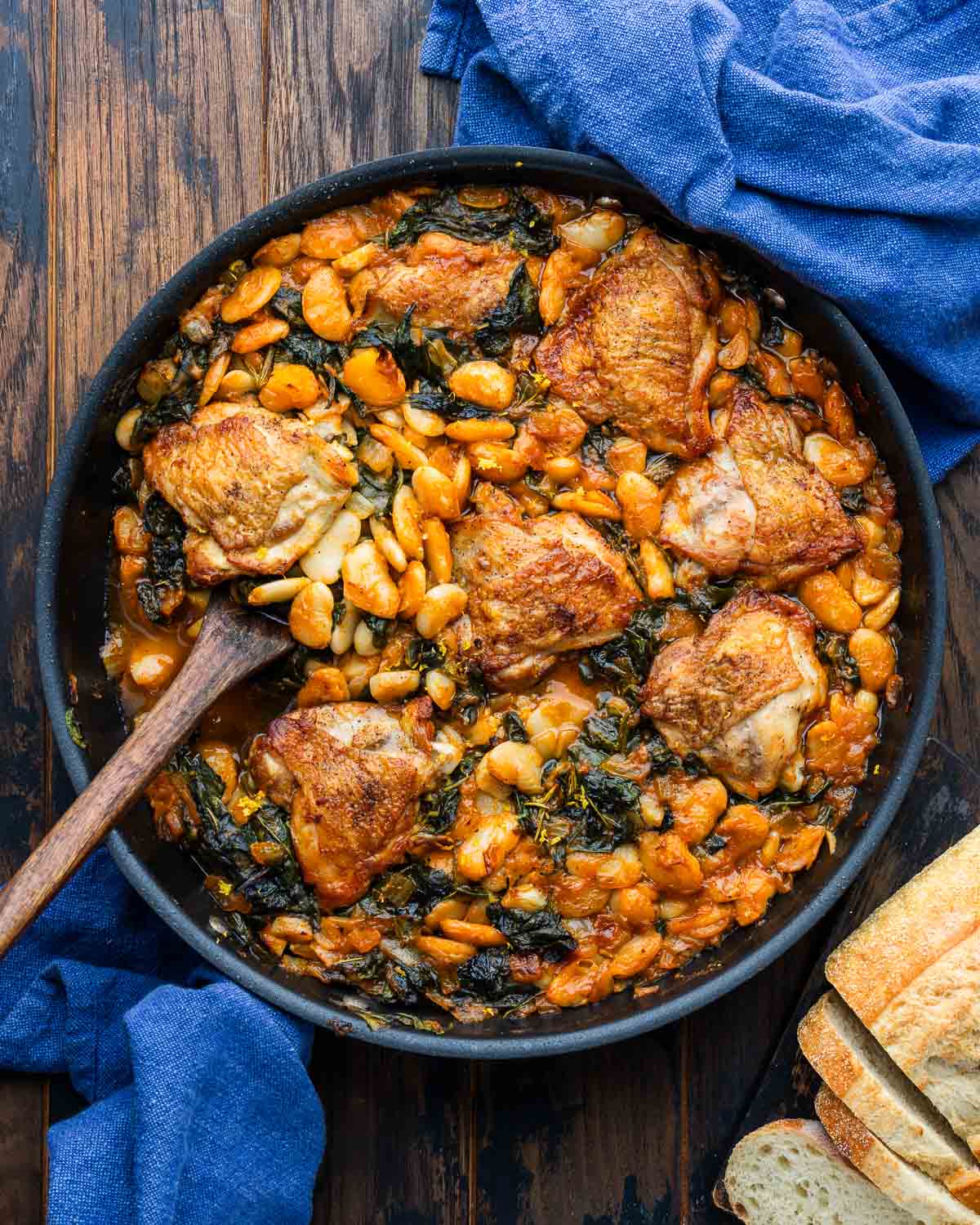 Chicken thighs with brothy beans in large pan with loaf of sliced bread on the side.