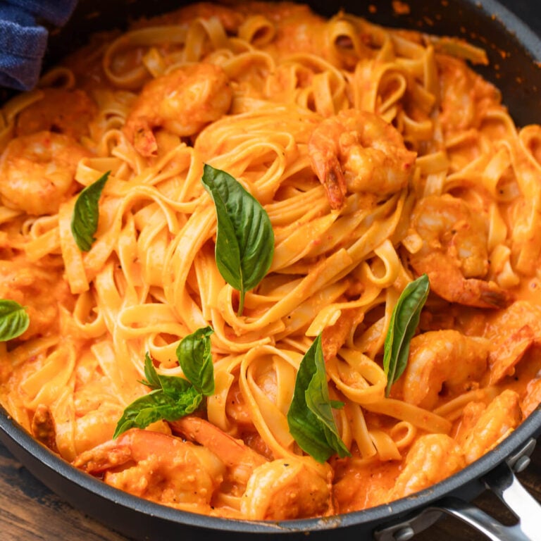 Roasted red pepper pasta with shrimp in black pan garnished with basil leaves.