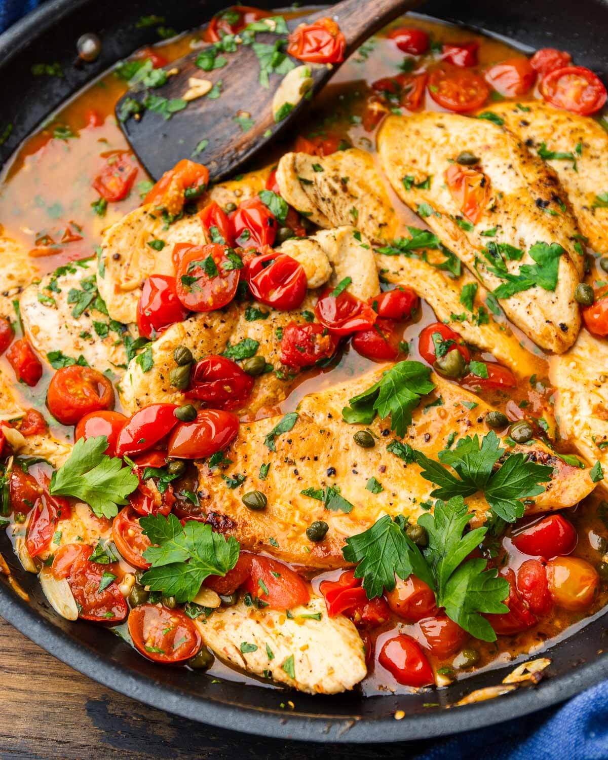 Chicken with capers and tomatoes in large black pan.