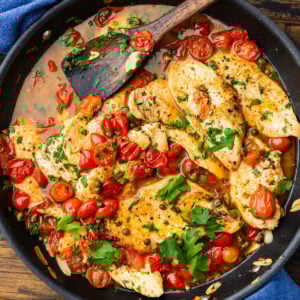 Large black pan of chicken with capers and tomatoes.