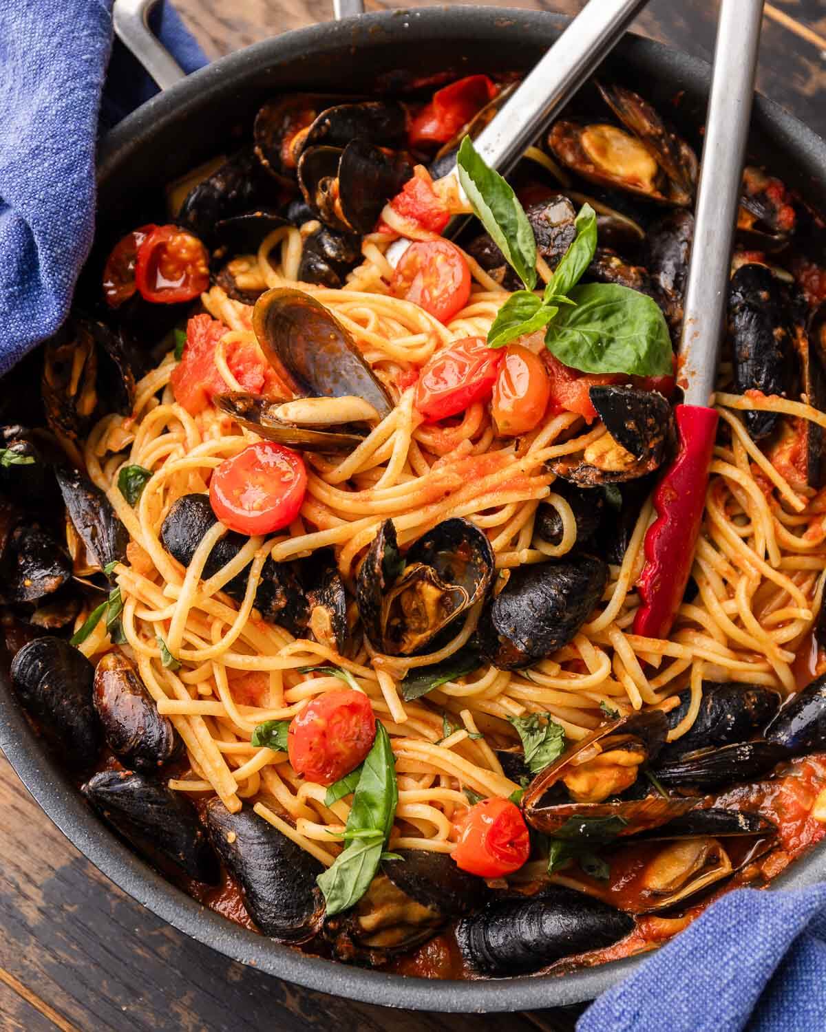 Linguine with mussels in large black pan with tongs.