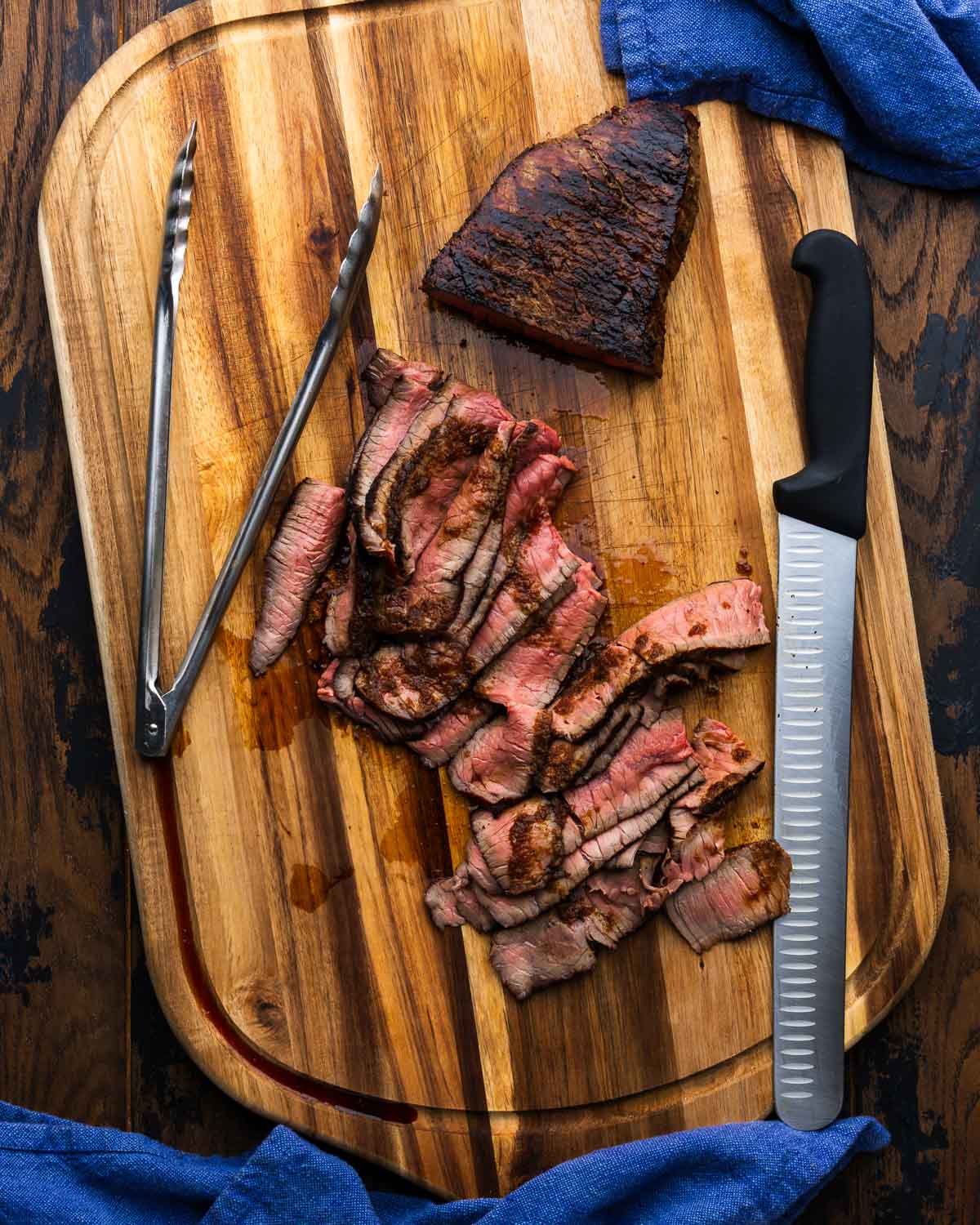 Sliced London Broil on cutting board with knife and tongs.