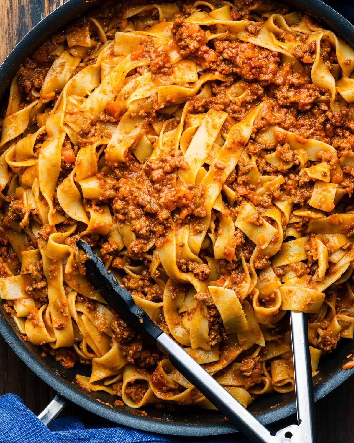 Pappardelle Bolognese in large pan with tongs.