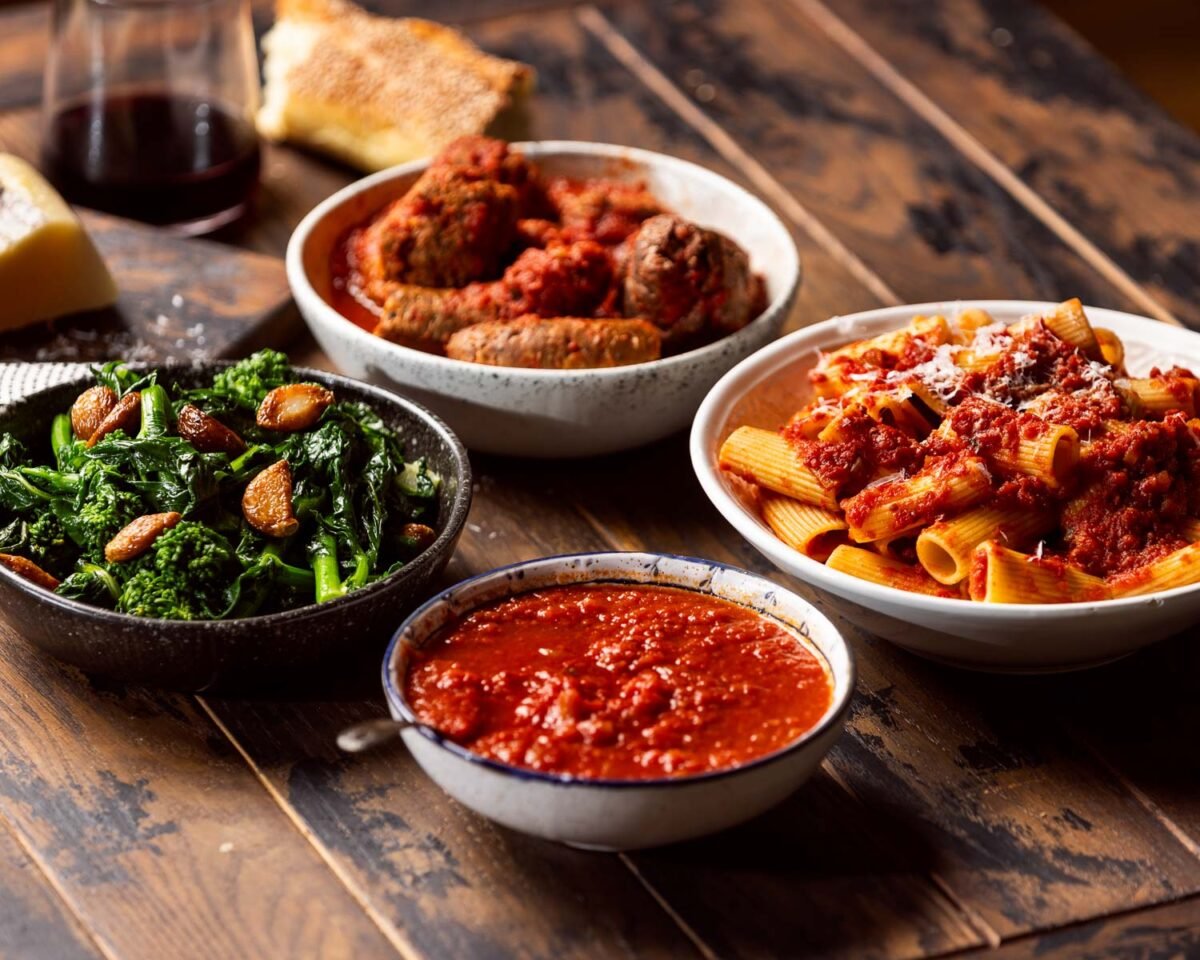 Pasta with Sunday sauce, bowl of sausage and meatballs, bowl of broccoli rabe, and glass of red wine.