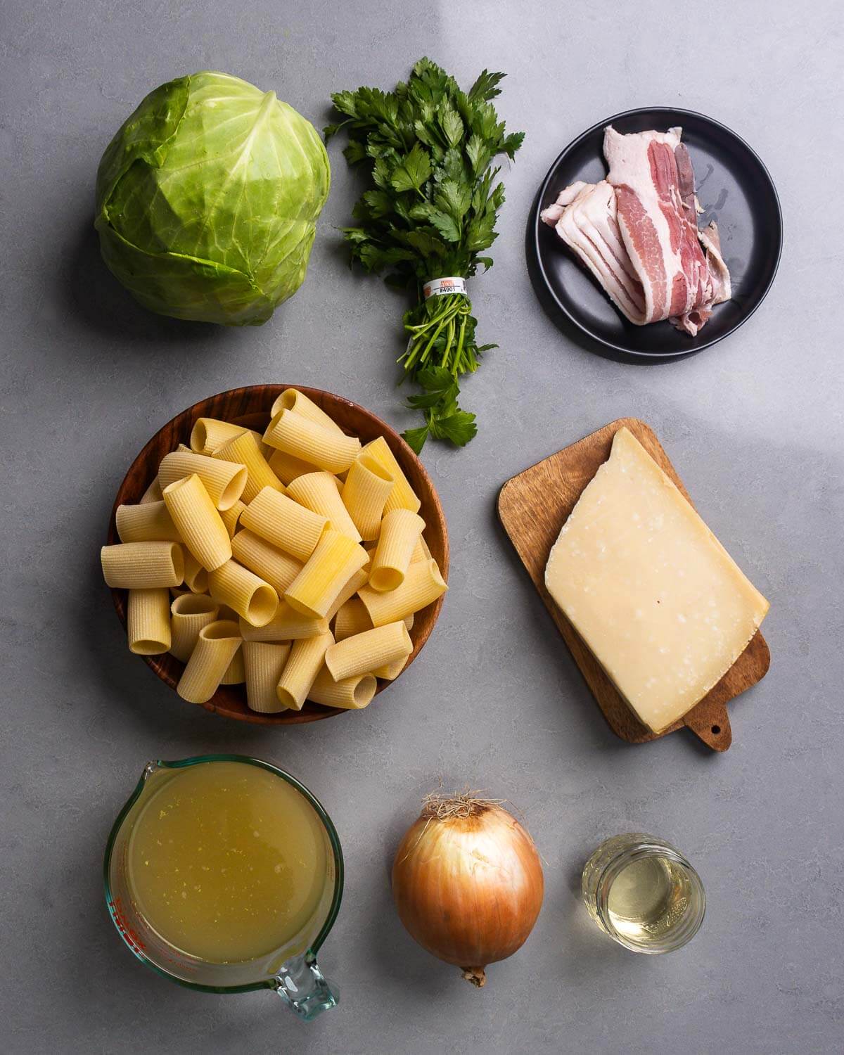 Ingredients shown: cabbage, parsley, bacon, rigatoni, block of parmesan, chicken stock, onion, and glass of white wine.