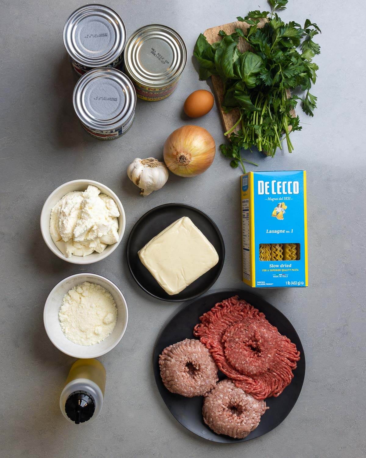 Ingredients shown: 3 cans of plum tomatoes, egg, basil, parsley, onion, garlic, ricotta, mozzarella, lasagna noodles, pecorino romano, olive oil, ground beef, and ground pork.