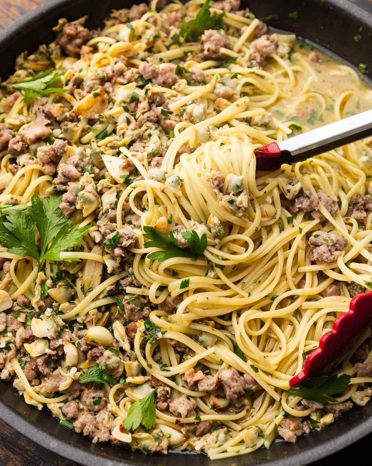 Linguine with sausage and clams in black pan with red tongs.