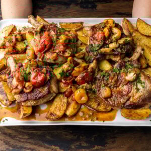 Hands holding platter of pork chops with potatoes and peppadew peppers.