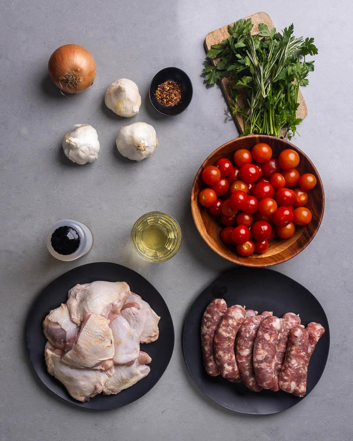Ingredients shown: onion, garlic, hot red pepper flakes, parsley, rosemary, cherry tomatoes, olive oil, white wine, chicken thighs, and Italian sausage.