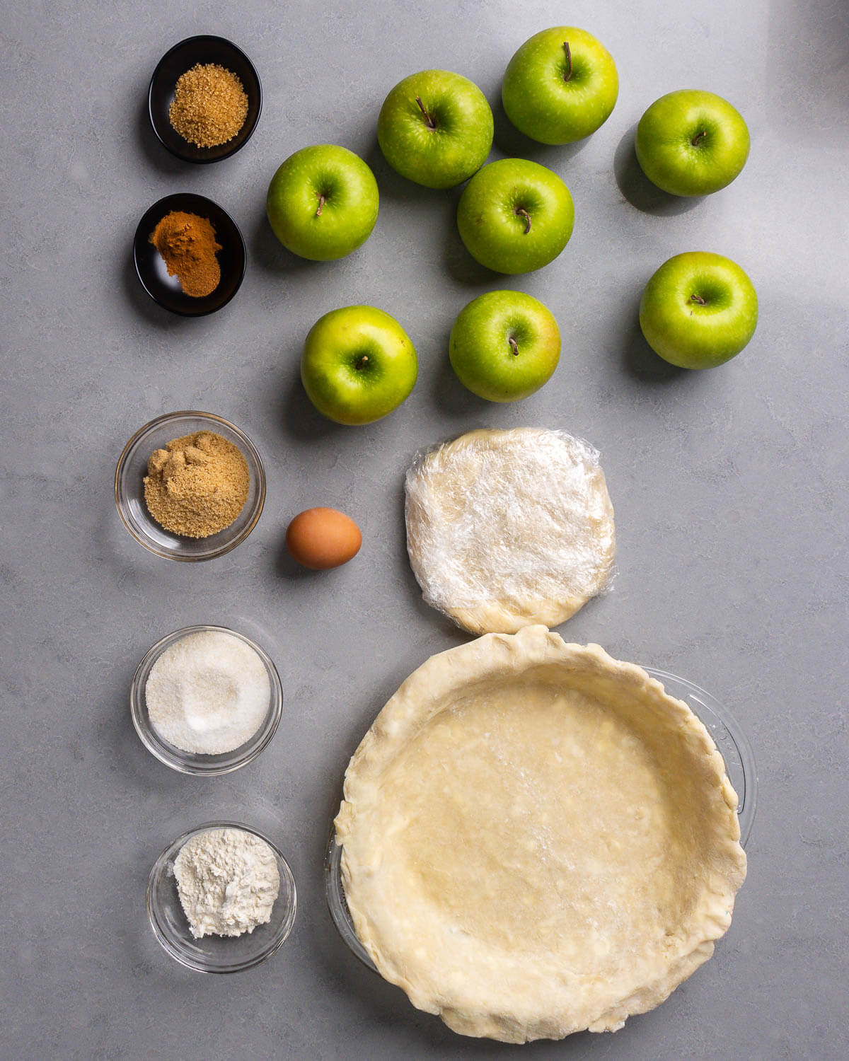 Apple pie ingredients shown: turbinado sugar, cinnamon, nutmeg, Granny Smith apples, egg, chilled pie crusts, brown sugar, sugar, and flour.