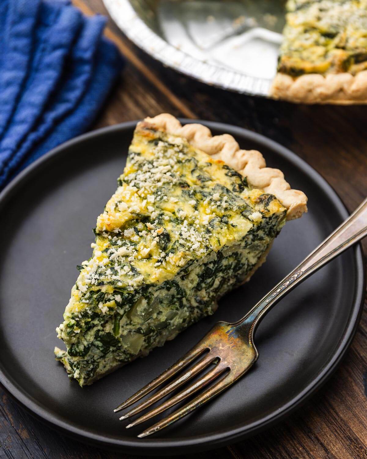 Slice of spinach ricotta pie on black plate with pie tin in the background.