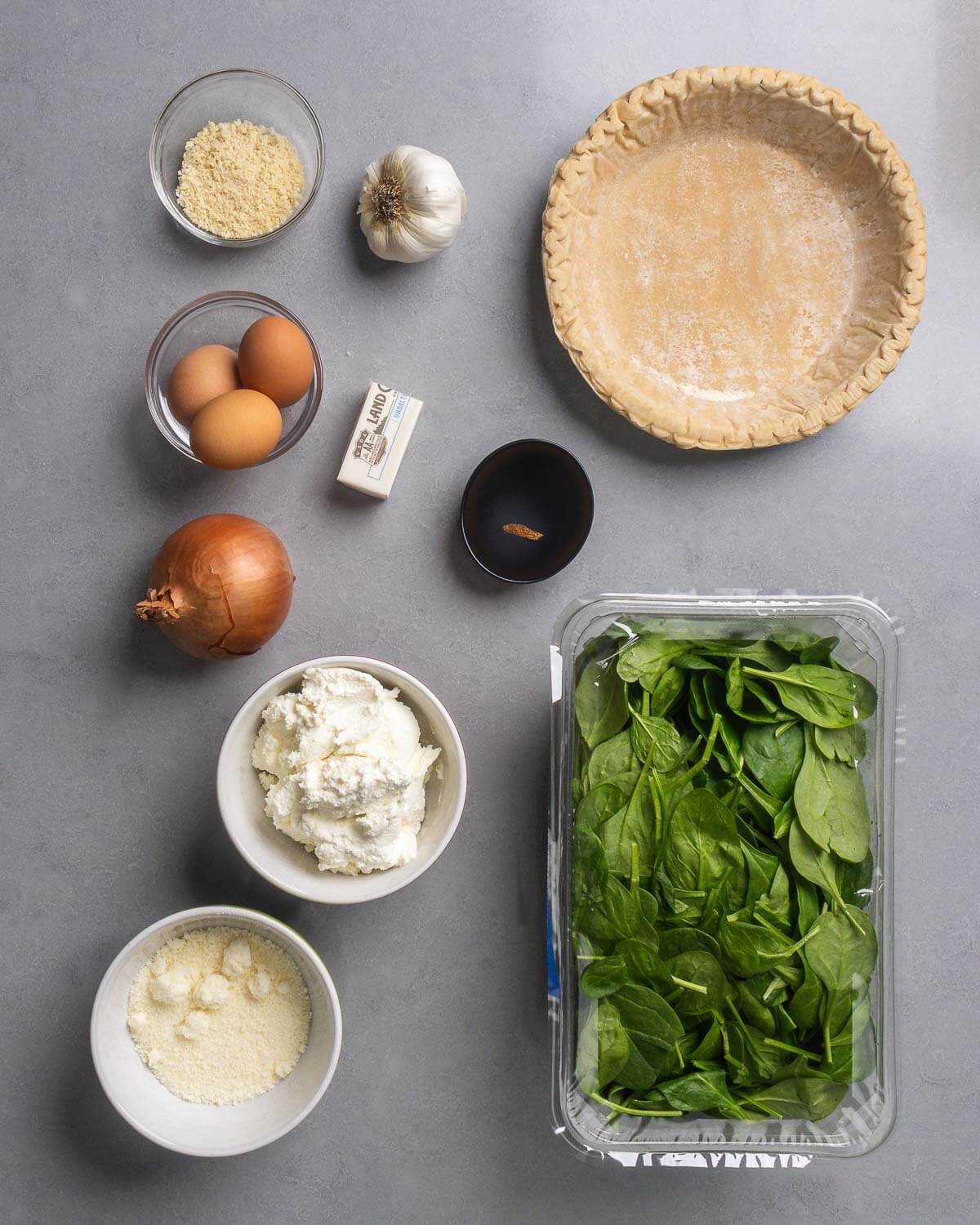 Ingredients shown: panko breadcrumbs, garlic, one 9-inch pie crust, eggs, butter, nutmeg, onion, ricotta, parmesan cheese, and baby spinach.