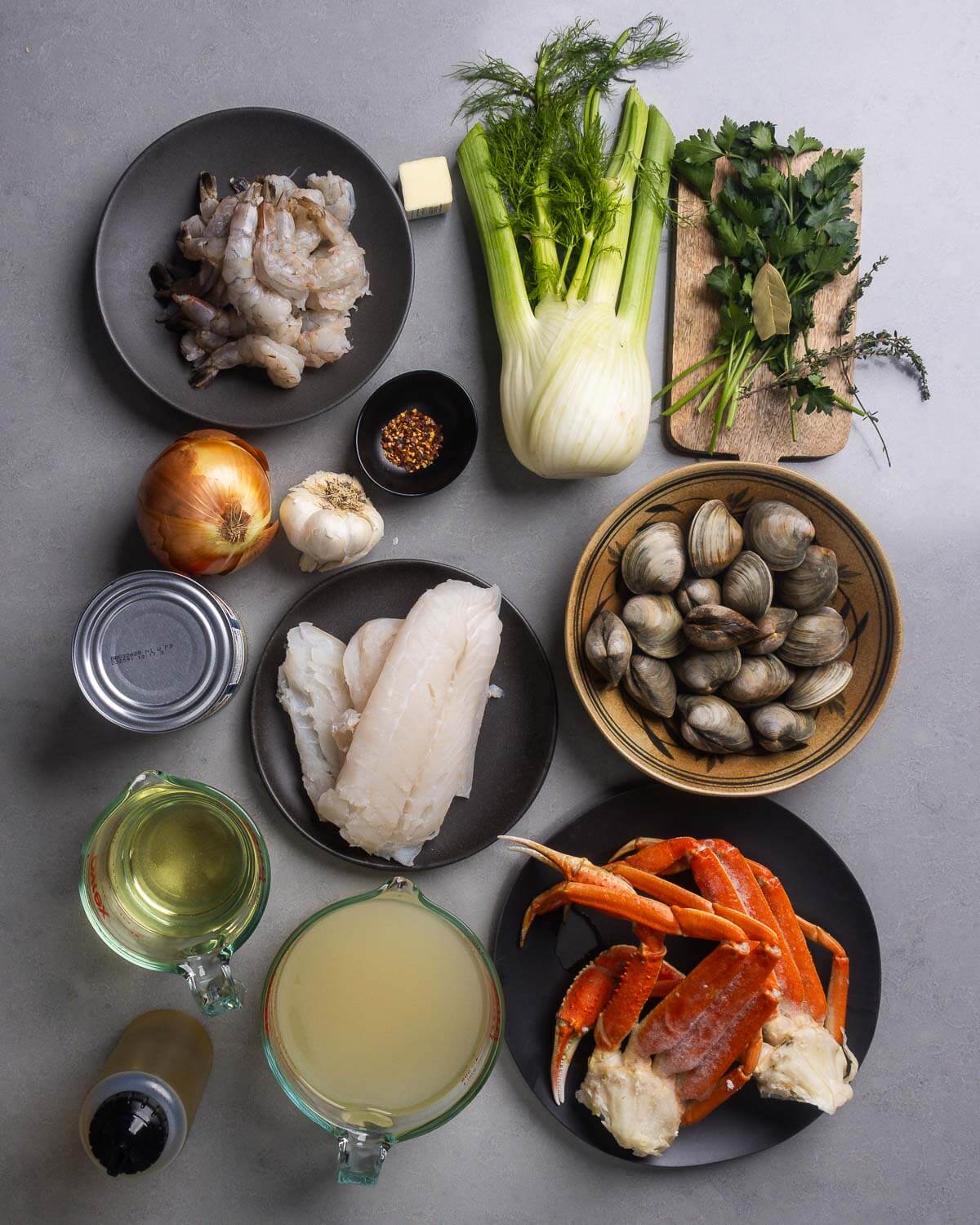Ingredients shown: shrimp, crushed red pepper flakes, fennel, parsley, bay leave, thyme, onion, garlic, canned tomatoes, cod, clams, wine, olive oil, clam juice, and crab legs.