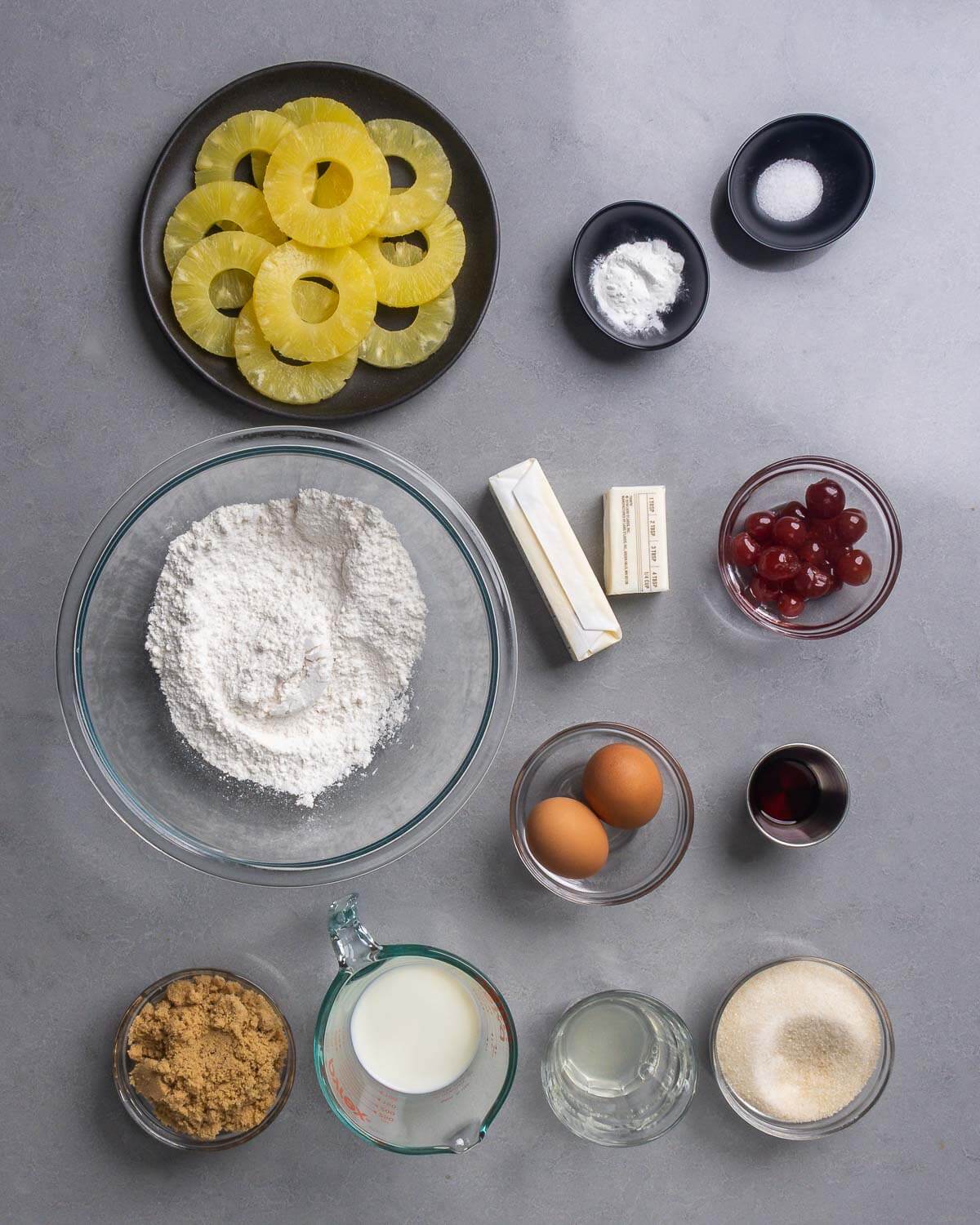 Ingredients shown: pineapple rings, baking powder, salt, flour, butter, cherries, eggs, vanilla extract, brown sugar, milk, pineapple juice, and granulated sugar.