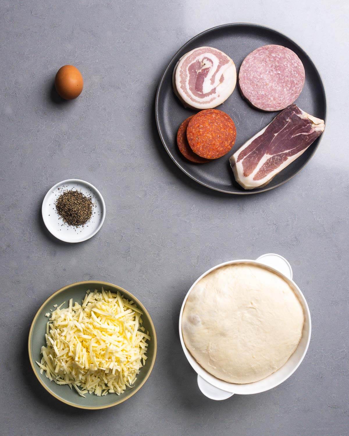 Ingredients shown: egg, pancetta, salami, pepperoni, prosciutto, black pepper, shredded provolone, and dough ball.