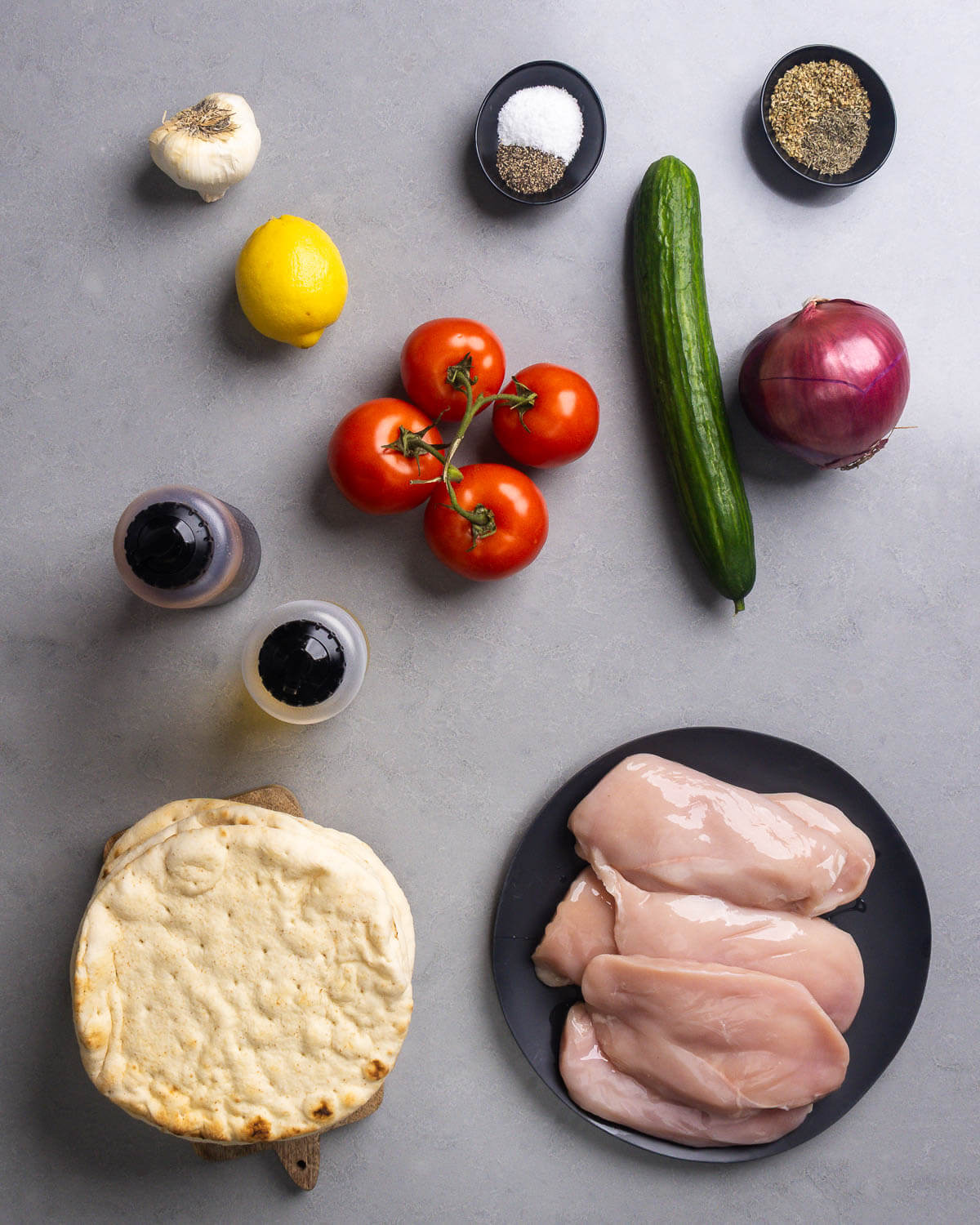 Ingredients shown: garlic, lemon, tomatoes, salt, pepper, cucumber, oregano, red onion, olive oil, red wine vinegar, pita, and chicken breasts.