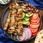 Grey plate with chicken souvlaki, kalamata olives, cucumbers, tomatoes, and red onion.
