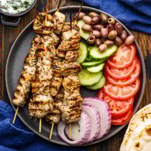 Grey plate with chicken souvlaki, kalamata olives, cucumbers, tomatoes, and red onion.