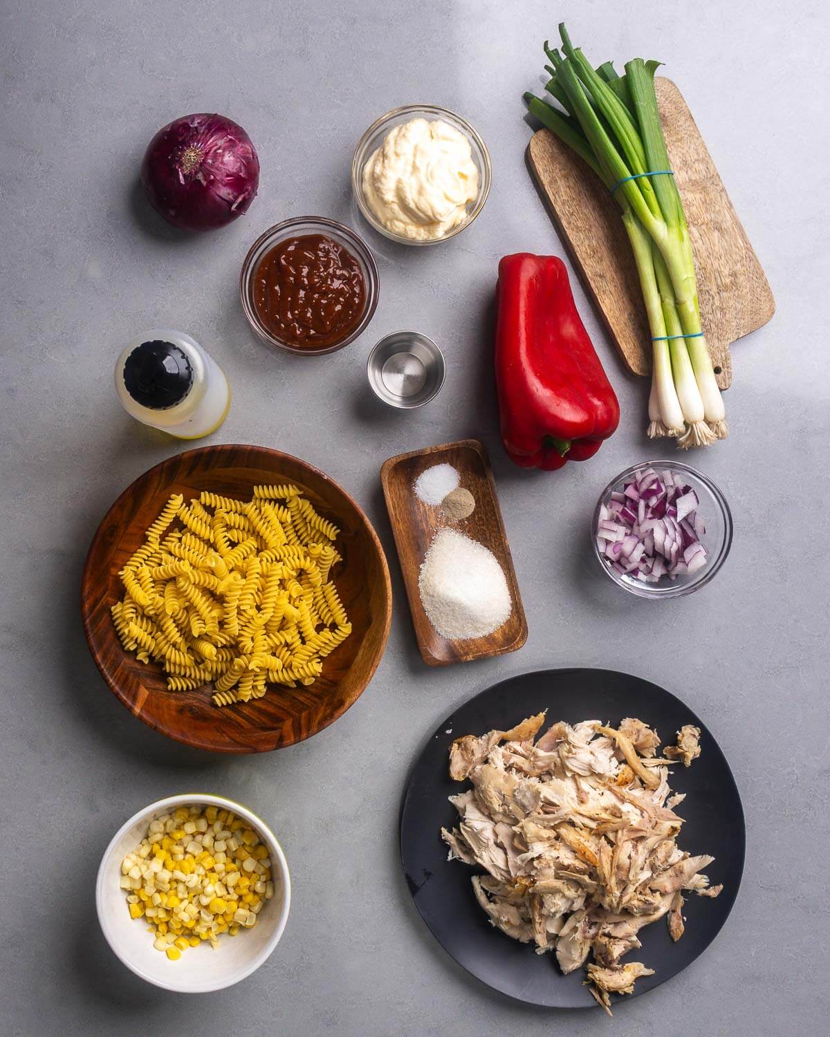 Ingredients shown: red onion, mayo, bbq sauce, vinegar, green onions, bell pepper, oil, pasta, spices, corn, and broken down rotisserie chicken.