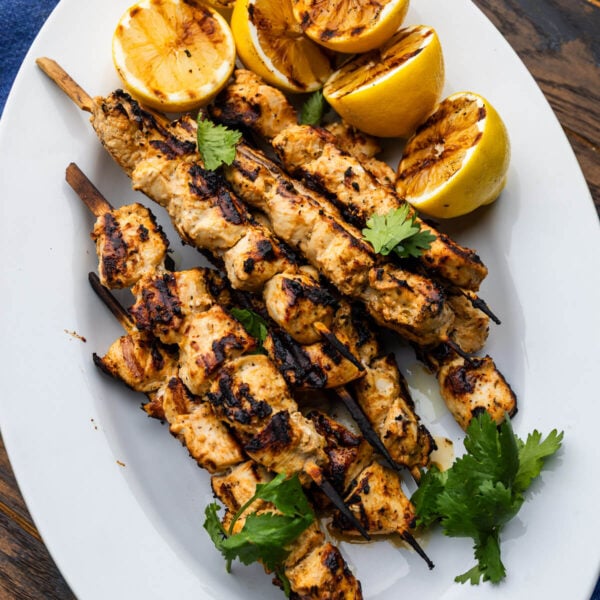 White platter with grilled lemon yogurt chicken kebabs topped with cilanto garnish and grilled lemons.