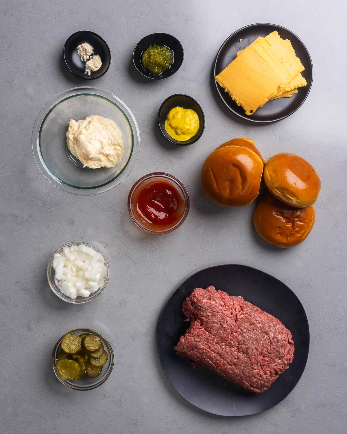 Ingredients shown: garlic and onion powder, relish, mustard, American cheese, mayonnaise, ketchup, brioche buns, diced onion, pickle chips, and ground chuck.