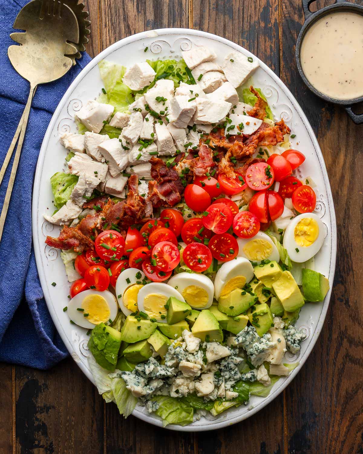 Cobb salad on white platter with spoons and extra dressing on the side.