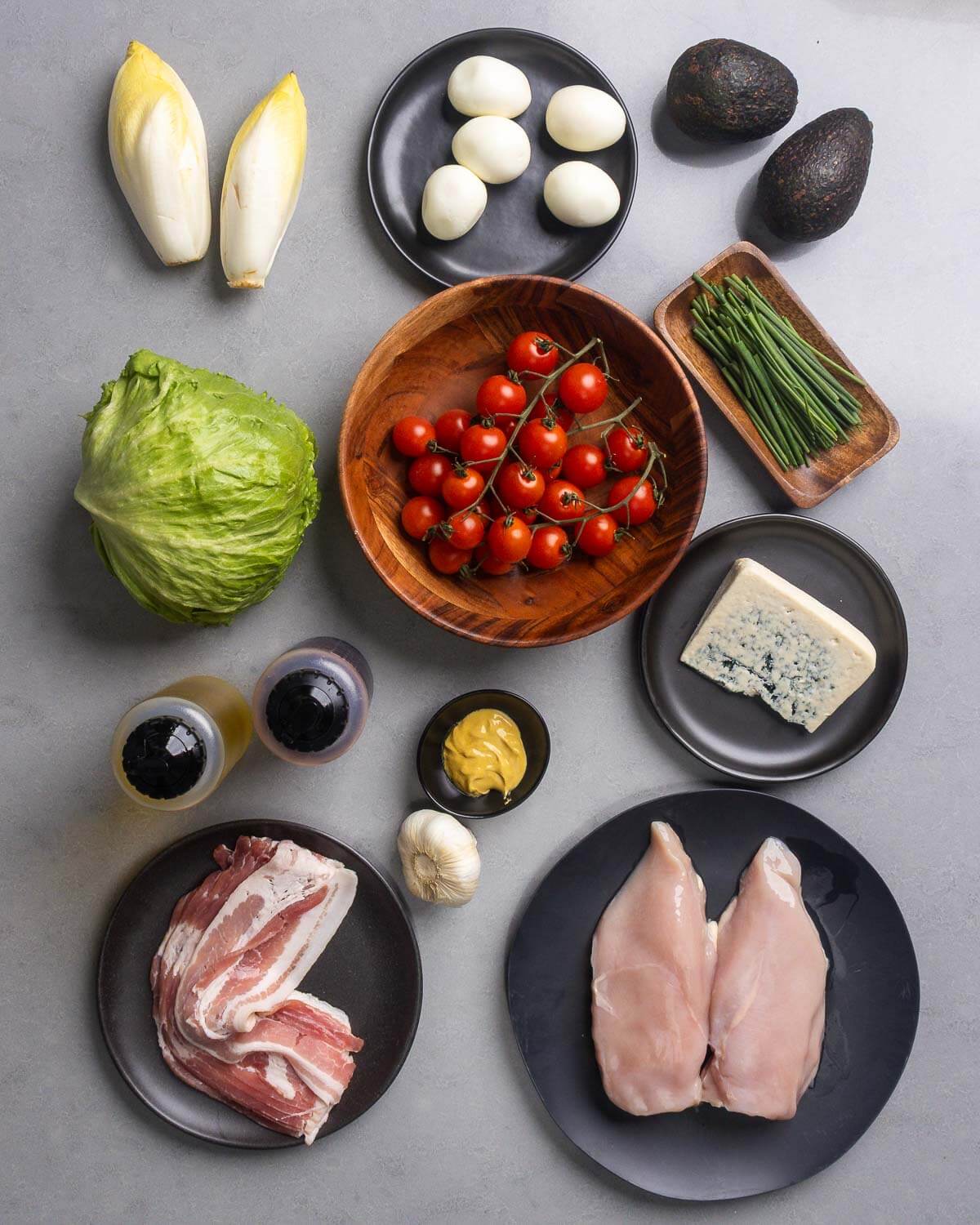 Ingredients shown: endive, eggs, avocados, lettuce, tomatoes, chives, blue cheese, olive oil, vinegar, dijon mustard, garlic, bacon, and chicken breasts.