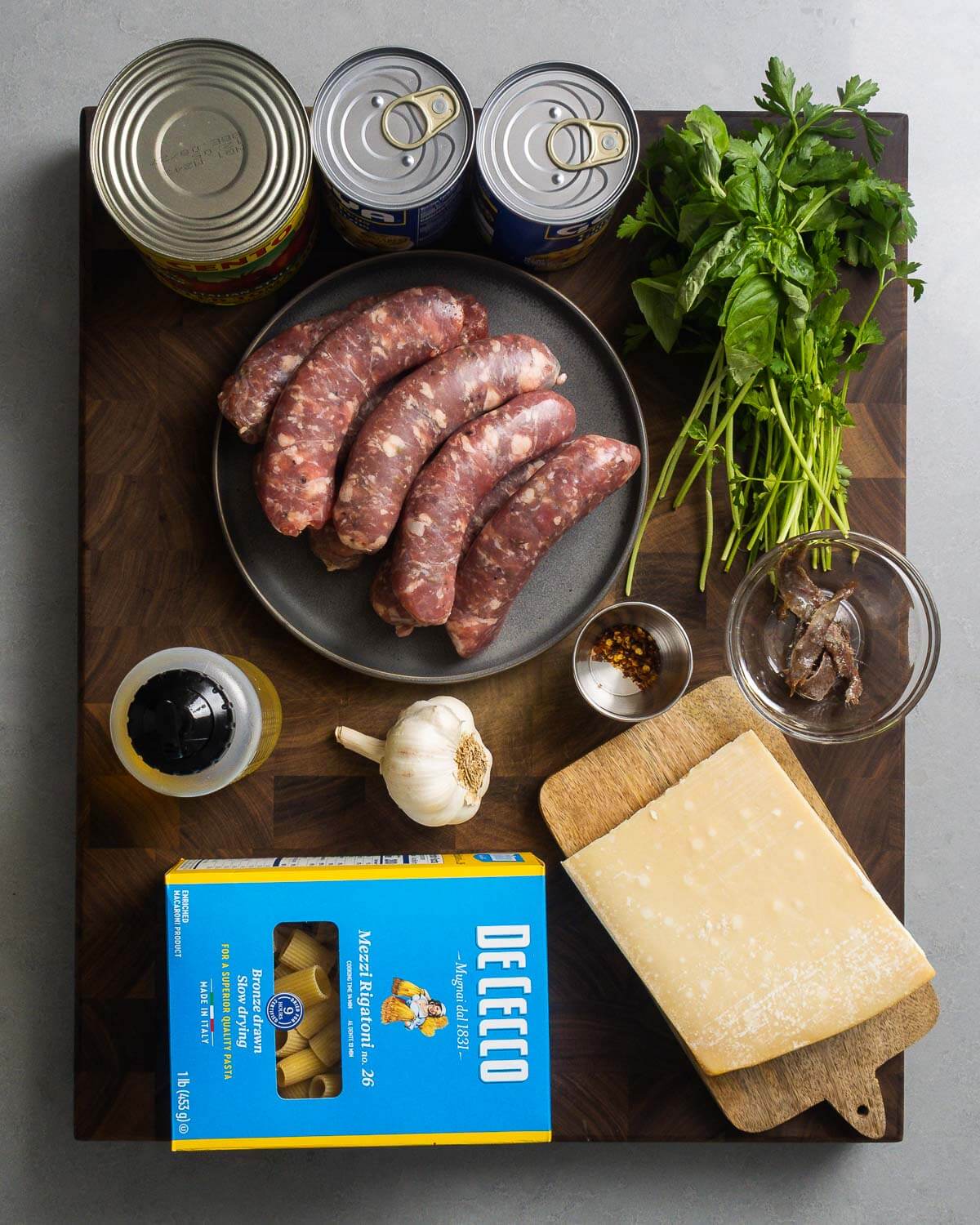 Ingredients shown: canned tomatoes, canned chickpeas, basil, parsley, Italian sausage, hot red pepper flakes, anchovies, olive oil, garlic, rigatoni pasta, and block of Parmigiano Reggiano.