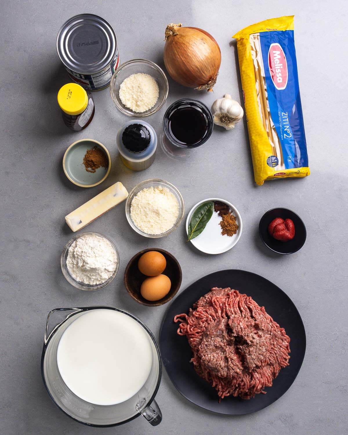 Ingredients shown: canned tomatoes, honey, parmesan cheese, onon, pastitsio pasta #2, red wine, garlic, olive oil, nutmeg, cinnamon, butter, bay leaves, tomato paste, flour, eggs, milk, and ground beef.